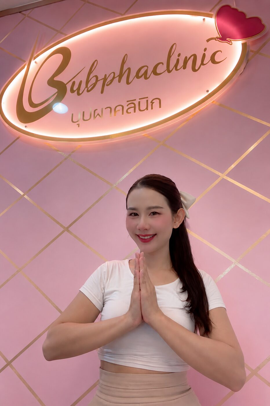 ผู้ดูแลบุปผาคลินิกยกมือไหว้ต้อนรับ หน้าป้ายไฟนีออนสีชมพู Bubphaclinic บุปผาคลินิก